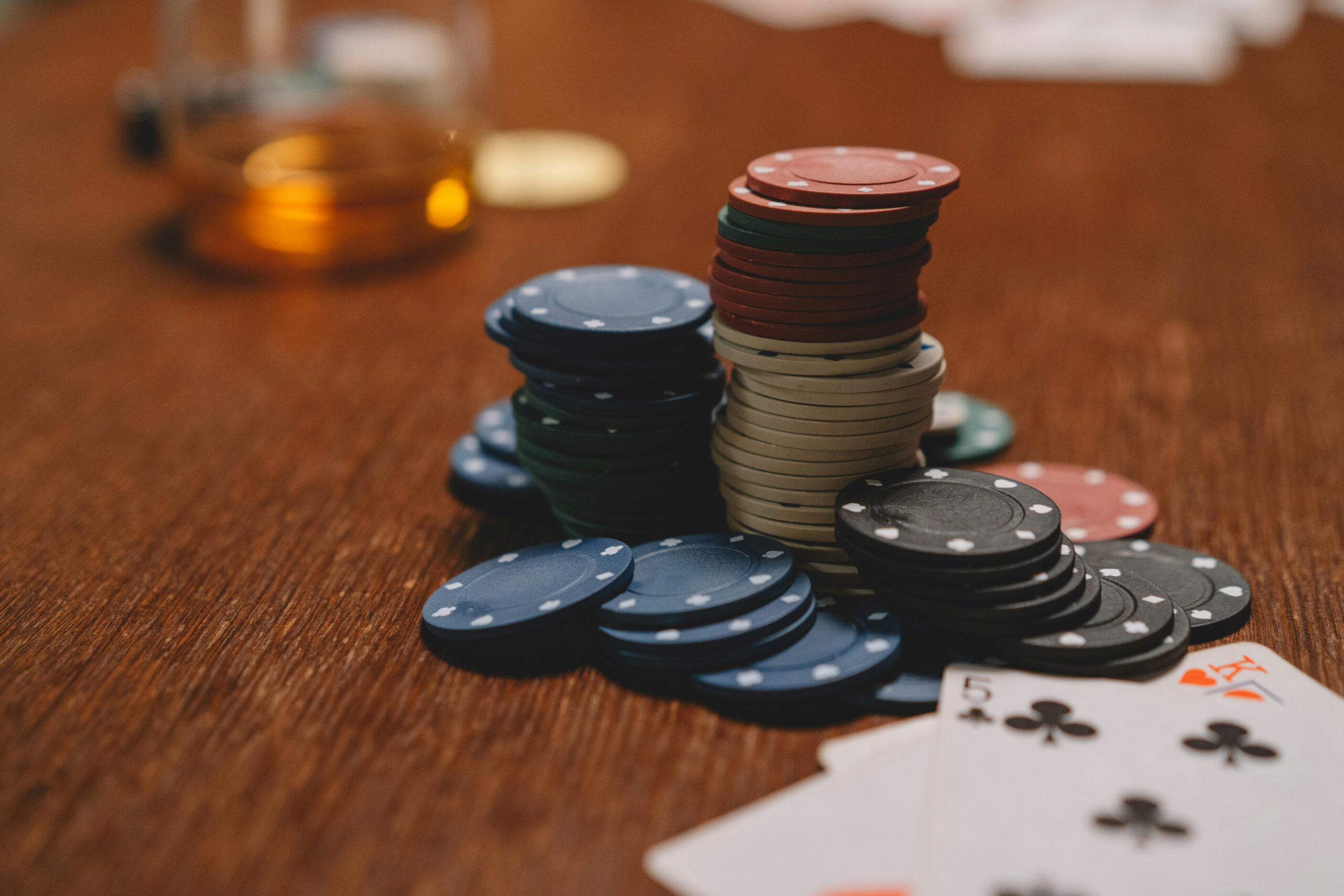 Poker chips stacked on a wooden table with playing cards and blurred glass in the background.